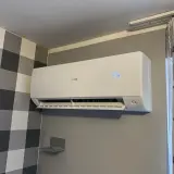 Biała klimatyzacja Haier zamontowana na szarej ścianie, obok zasłony.