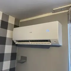 Biała klimatyzacja Haier zamontowana na szarej ścianie, obok zasłony.